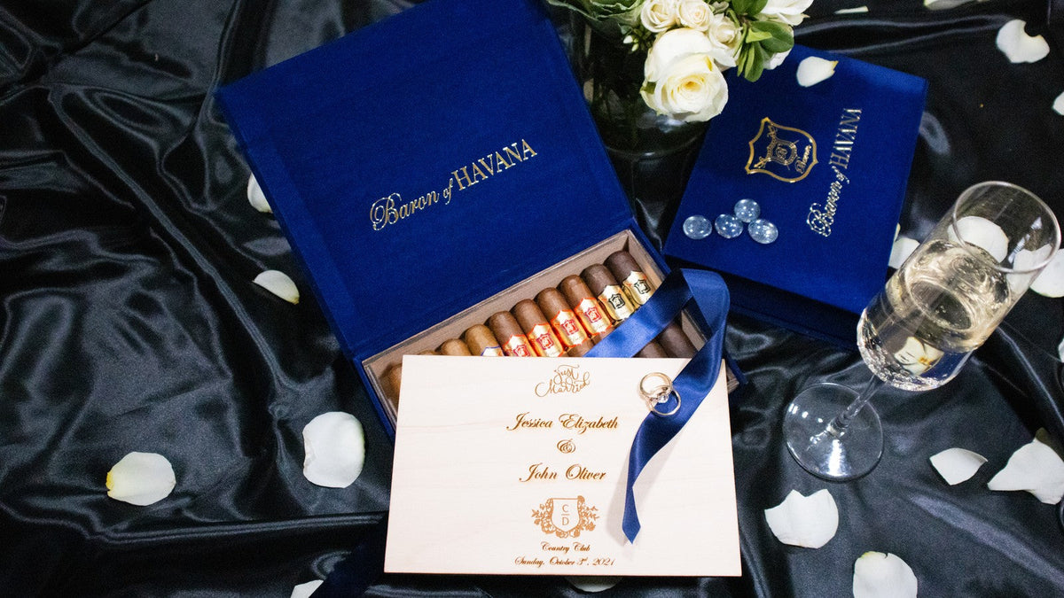 An open blue velvet Baron of HAVANA box of cigars with an engraved card, white roses and champagne on black satin