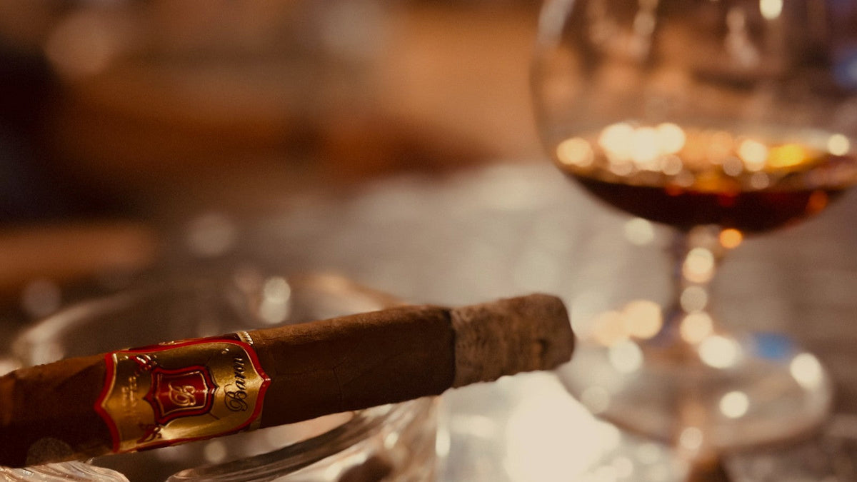 A banded Baron of HAVANA cigar resting on a crystal ashtray beside a snifter of cognac in warm evening light