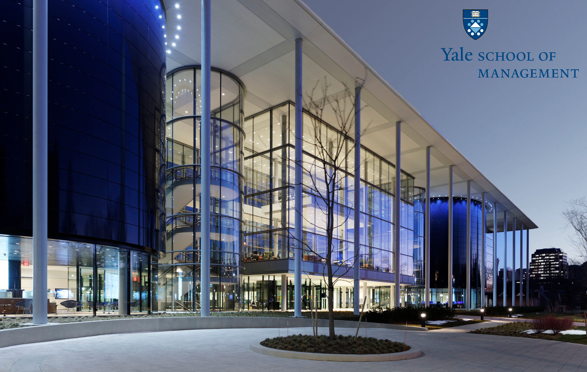 The Yale School of Management building at dusk, its glass front lit from inside.